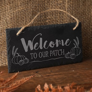 May include: Black slate welcome sign with white text that reads "Welcome to our patch". The sign is decorated with a white outline of pumpkins and leaves.
