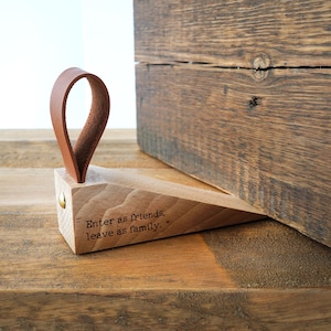 May include: A wooden doorstop with a brown leather loop handle. The doorstop is wedge-shaped and has the engraved text "Enter as friends, leave as family." A wooden block is in the background.