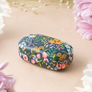 Könnte beinhalten: Ein Brillenetui mit floralem Muster auf dunkelblauem Hintergrund und rosa, gelben und weißen Blumen. Das Etui ist oval geformt und hat einen Klappdeckel.