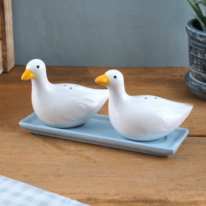 May include: A white ceramic duck-shaped salt and pepper set, featuring yellow beaks and black eyes. The ducks are presented on a light blue rectangular tray. The set is on a wooden surface.