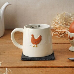 Könnte beinhalten: Cremefarbene Keramik-Tasse mit braunem Hühnerdesign und dem Schriftzug "Rise & Shine" im Rand. Die Tasse steht auf einem schwarzen quadratischen Untersetzer. Eine Teekanne und ein Eierbecher sind im Hintergrund.