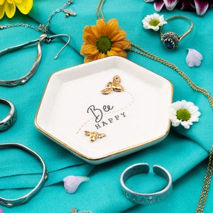 Könnte beinhalten: Eine weiße sechseckige Keramik-Schmuckschale mit goldenem Rand. Die Schale hat ein goldenes Bienen-Design und den Text "Bee Happy". Die Schale ist von Schmuck, Blumen und Stoff umgeben.