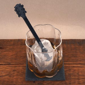 May include: A clear glass with a guitar-shaped ice cube and a black guitar-shaped stirrer. The glass sits on a black coaster on a wooden surface. The ice cube is in the shape of a guitar.