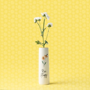Puede incluir: Jarrón de cerámica blanco con un diseño de abeja y flores. El jarrón tiene el texto "Bee Happy" en la parte delantera. Un ramo de flores blancas está en el jarrón.