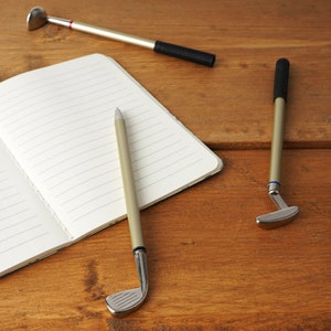 Könnte beinhalten: Drei Stifte in Form von Golfschlägern auf einer Holzoberfläche. Zwei der Stifte sind silberfarben und einer ist schwarz. Die Stifte liegen neben einem leeren Notizbuch.