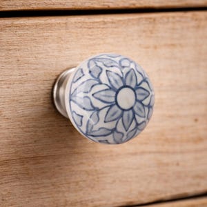May include: A ceramic drawer knob featuring a blue and white floral pattern. The knob is round and mounted on a silver-coloured metal base. It is set against a light-coloured wooden drawer. The floral design is a central feature.