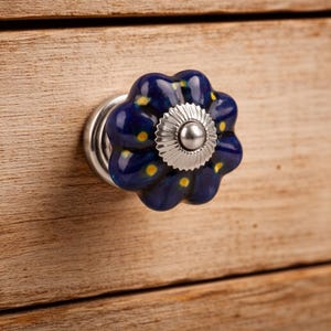 May include: A flower-shaped ceramic drawer knob in a deep blue color with small yellow dots. The center of the knob features a circular silver-toned metal plate with a ridged edge and a smooth, rounded silver-toned metal button in the middle.
