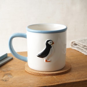 May include: White ceramic mug featuring a 3D puffin design. The mug has a light blue handle and rim, and rests on a cork coaster. The puffin is black and white with an orange beak and feet. A decorative item for the home.