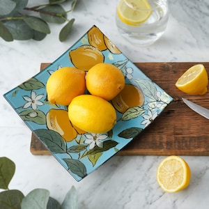 May include: A blue ceramic serving dish with a lemon motif. The dish is filled with four lemons, and there are white flowers and green leaves painted on the dish.