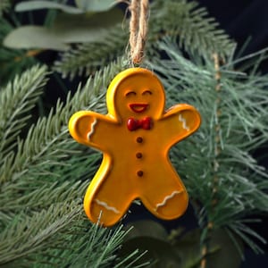 Peut inclure: Ornement de Noël en forme de bonhomme en pain d'épices. L'ornement jaune présente des détails de glaçage blanc, un nœud papillon rouge et un visage souriant. Il est suspendu par une ficelle en corde naturelle sur un fond de branches de conifères.