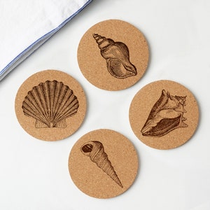May include: Four round cork coasters featuring detailed illustrations of seashells. The coasters are a natural tan colour, and the seashell designs are in a dark brown. A white and blue striped cloth is in the background.