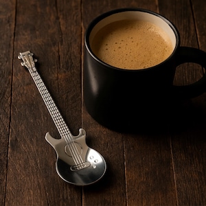 Puede incluir: Una taza de café negra llena de café se encuentra junto a una cuchara plateada con forma de guitarra sobre una superficie de madera oscura. La cuchara tiene un diseño detallado, que se asemeja a una guitarra eléctrica en miniatura. El café es marrón claro con espuma.