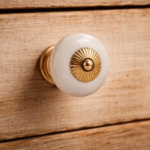 May include: A round white ceramic cabinet knob featuring a central gold-toned metal accent with a sunburst-like fluted design and a small spherical center point, mounted on a gold-toned metal base attached to a light-colored wooden surface.