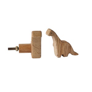 Puede incluir: Dos pomos de madera con forma de dinosaurio. Un pomo es de forma rectangular simple, el otro tiene forma de dinosaurio. Ambos pomos son de madera marrón claro.