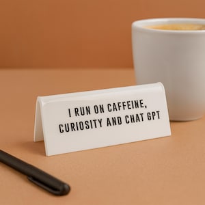 May include: A white desk sign with the text "I RUN ON CAFFEINE, CURIOSITY AND CHAT GPT" in black. A white mug and a black pen are also in the image. The background is a solid tan colour.