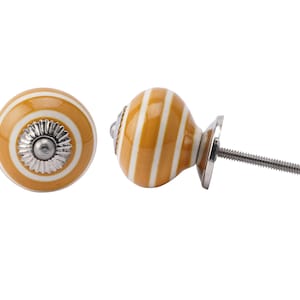 Peut inclure: Deux boutons de meuble décoratifs. Chaque bouton présente un motif rayé jaune moutarde et blanc. Les boutons ont une base et une vis en métal argenté pour l'installation. L'un des boutons est présenté de face, l'autre de côté.