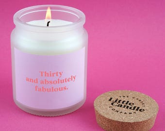 Może przedstawiać: Zapalone biała świeca w mrożonym szklanym słoiku z różową etykietą z napisem "Thirty and absolutely fabulous". Korek z napisem "The Cheeky Little Candle Company" leży obok słoika. Tło jest jednolicie różowe.