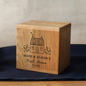 May include: A wooden savings box with a slot on top. The box is engraved with a house illustration and the text "MARK & SARAH'S First Home FUND". The box is made of light brown wood and sits on a dark blue surface.