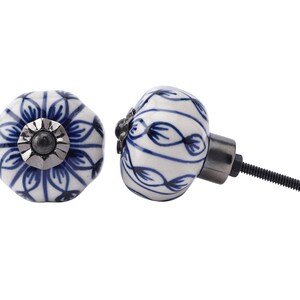 Pode incluir: Dois botões de armário de cerâmica com fundo branco e desenhos florais azuis. Um botão tem um acento de metal prateado no centro. O outro botão tem um parafuso de metal preto.