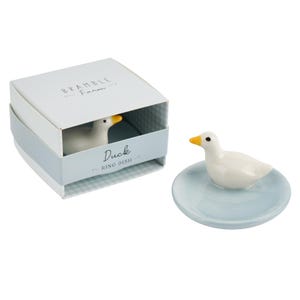 May include: A white ceramic duck ring dish with a yellow beak and black eyes. The duck sits on a light blue dish. The dish is next to a box with the words "Bramble Farm" and "Duck Ring Dish".
