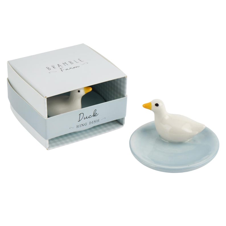 May include: A white ceramic duck ring dish with a yellow beak and black eyes. The duck sits on a light blue dish. The dish is next to a box with the words "Bramble Farm" and "Duck Ring Dish".