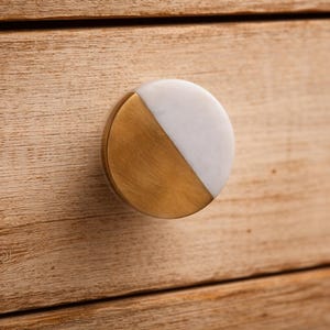 May include: A circular cabinet knob featuring a split design with one half composed of brushed gold-tone metal and the other half made of smooth white stone. The knob is mounted onto a light-coloured wooden surface with a visible horizontal grain pattern.