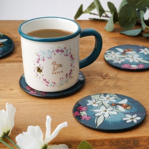 May include: A white ceramic mug with a teal rim and handle. The mug has a floral design with a bee and the text "Bee Happy". The mug is sitting on a blue and white floral coaster.