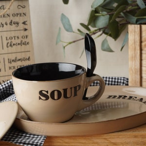 Puede incluir: Un tazón de sopa de cerámica beige con un interior negro y la palabra "SOUP" impresa en el lateral. El tazón está sentado en una bandeja de servir de cerámica beige con la palabra "BREAD" impresa en el lateral. La bandeja está sobre un mantel a cuadros blanco y negro.