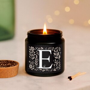 Puede incluir: Una vela negra encendida en un tarro con la letra "E" blanca, rodeada de dise&ntilde;os florales. La vela est&aacute; sobre una superficie de m&aacute;rmol, con una tapa de corcho y cerillas cerca. La llama de la vela brilla.
