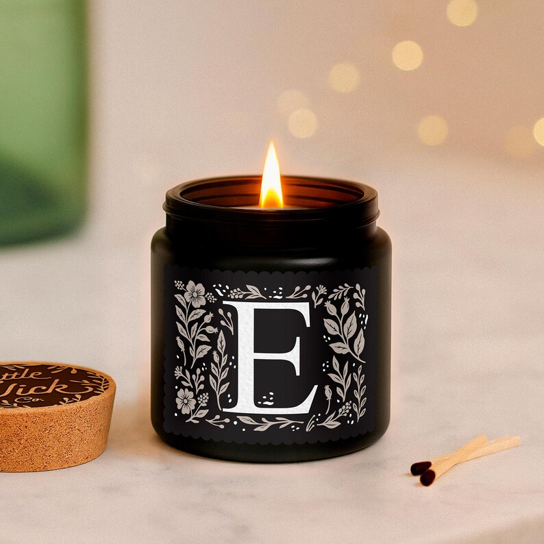Puede incluir: Una vela negra encendida en un tarro con la letra "E" blanca, rodeada de dise&ntilde;os florales. La vela est&aacute; sobre una superficie de m&aacute;rmol, con una tapa de corcho y cerillas cerca. La llama de la vela brilla.