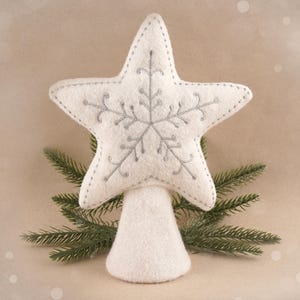 Könnte beinhalten: Ein Weihnachtsbaumschmuck aus weißem Filz in Form eines Sterns mit einem silbernen Schneeflockenmuster. Der Stern befindet sich auf einer weißen, kegelförmigen Basis, mit grünen Tannenzweigen.
