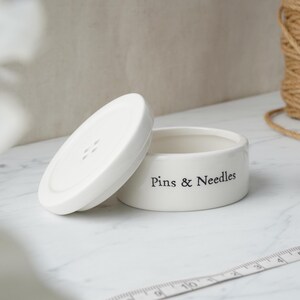 May include: White ceramic button-shaped container with a lid. The container has the words "Pins & Needles" printed on the side.