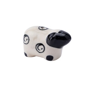 May include: A ceramic sheep figurine with a white body, black head, and black spots. The sheep has decorative black swirl patterns on its body. The figurine is a decorative item.
