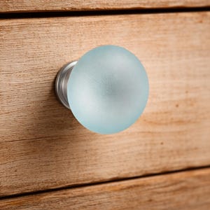 Könnte beinhalten: Ein kugelförmiger Möbelknopf mit einer mattierten hellblauen Glasoberfläche und einem runden silberfarbenen Metallsockel. Der Knopf ist an einer hellbraunen Holzschubladenfront mit sichtbarer horizontaler Holzmaserung und Fuge befestigt.