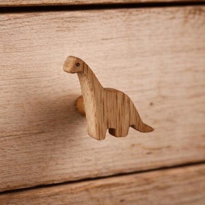 May include: A light brown wooden drawer knob shaped like a long-necked dinosaur. The knob features a visible wood grain pattern and a small dark dot representing the eye, attached to a light-colored wooden drawer front with a horizontal grain.