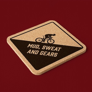 Puede incluir: Un posavasos de corcho con un diseño en blanco y negro. La mitad superior es de color marrón claro y la mitad inferior es negra. Un ciclista en bicicleta está en la mitad superior. Las palabras "MUD, SWEAT AND GEARS" están en blanco.