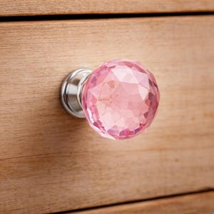 May include: A pink crystal cabinet knob with a faceted design and a silver base is attached to a wooden surface. The knob reflects light. The wooden surface has visible grain.