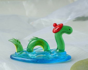 Könnte beinhalten: Eine grüne Glasfigur des Seeungeheuers von Loch Ness, das einen roten und schwarzen Tam o' Shanter trägt und in blauem Wasser schwimmt.