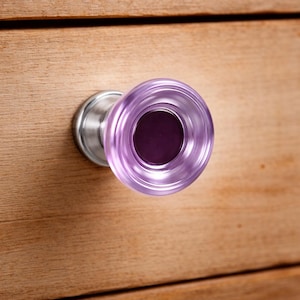 May include: A close-up of a cabinet knob. The knob features a transparent, light purple glass with a dark purple centre. The knob is mounted on a silver metal base and attached to a wooden surface.