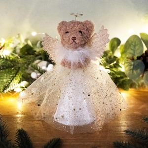 May include: A brown teddy bear angel tree topper with a white tulle skirt and gold glitter accents. The angel has a halo and feathery wings.