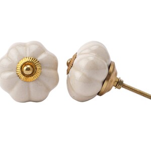 Pode incluir: Dois botões de armário de cerâmica branca em forma de abóbora com detalhes dourados. Os botões têm um acessório de parafuso.