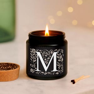 Puede incluir: Una vela negra encendida en un tarro con la letra blanca &laquo;M&raquo; rodeada de dise&ntilde;os florales. La vela est&aacute; sobre una superficie blanca, con un corcho y cerillas. La llama es brillante.