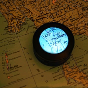 May include: A black compass with a blue illuminated screen showing a map of Phetchabun, Thailand, set against a vintage map. The compass is circular and features the text "Baten 6798 Khao Y Phetchabu Palaw H".