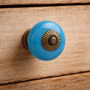 May include: A round, bright blue ceramic cabinet knob with a smooth, glossy finish. The center of the knob features a decorative, scalloped antique brass metal plate with a small, raised circular stud in the middle, attached to a brass base.