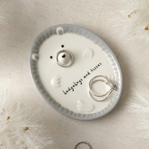 May include: White ceramic ring dish with a grey rim. The dish features a cartoon bear face with the text "hedgehugs and kisses".