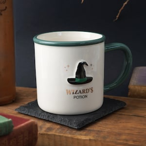 May include: White ceramic mug with a green rim and handle. The mug features a 3D witch's hat design and the text "Wizard's Potion" in gold lettering.