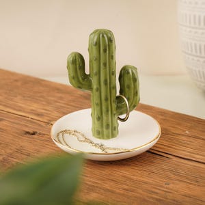 May include: A ceramic cactus-shaped jewelry holder. The green cactus sits atop a white dish with a gold rim. A gold ring is displayed on the cactus, and a gold necklace rests in the dish. The holder is on a wooden surface.