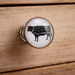 May include: A cabinet knob with a clear, round glass top featuring a black silhouette of a cow with labeled cuts of meat. The knob has a metallic bronze-colored rim and is mounted on a light brown wooden drawer.