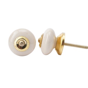 May include: Two off-white ceramic cabinet knobs with gold-colored metal accents. The knobs have a round shape with a decorative gold ring around the center. The screw is visible on the back of one knob. These knobs are suitable for furniture.