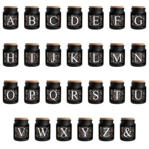 Puede incluir: Un conjunto de peque&ntilde;os frascos de vidrio negro con tapas de corcho, cada uno con una letra diferente del alfabeto en una fuente floral blanca. Los frascos est&aacute;n dispuestos en filas, ideales para regalos personalizados o almacenamiento.